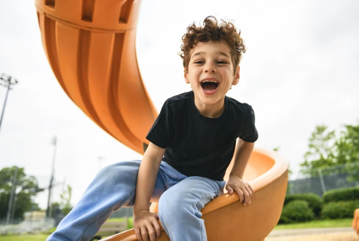 Un enfant souriant, vêtu d'un t-shirt noir et d'un pantalon bleu, est assis au bout d'un toboggan de couleur orange vif dans une aire de jeux.